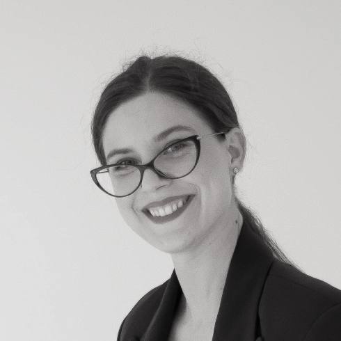 Tina Jura zâmbind, în sacou închis la culoare, pe un fundal deschis.